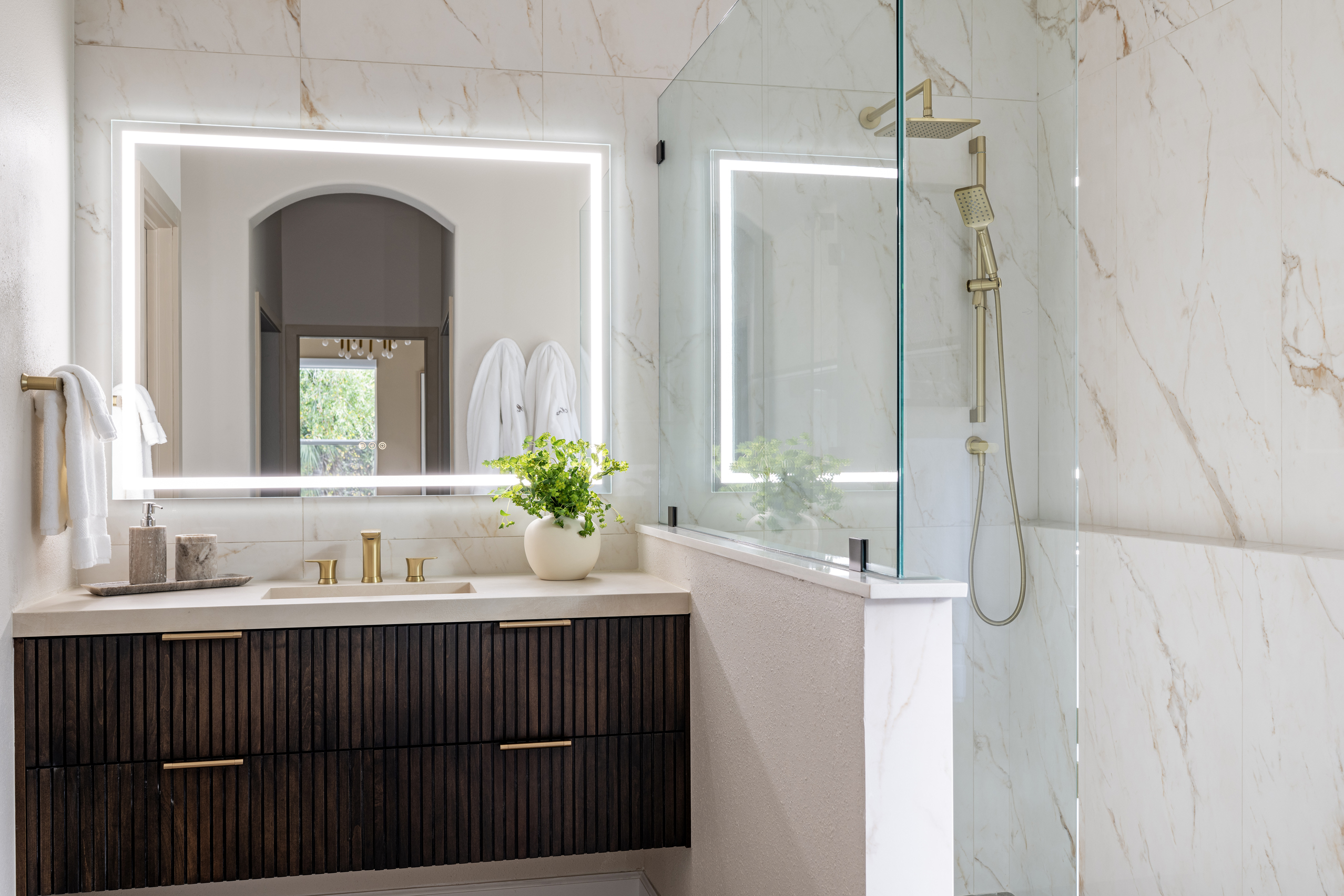 Luxury primary bathroom with full marble tile walls, dark fluted wood vanity, gold fixtures, backlit mirror, and a glass-enclosed walk-in shower designed RDM Interiors