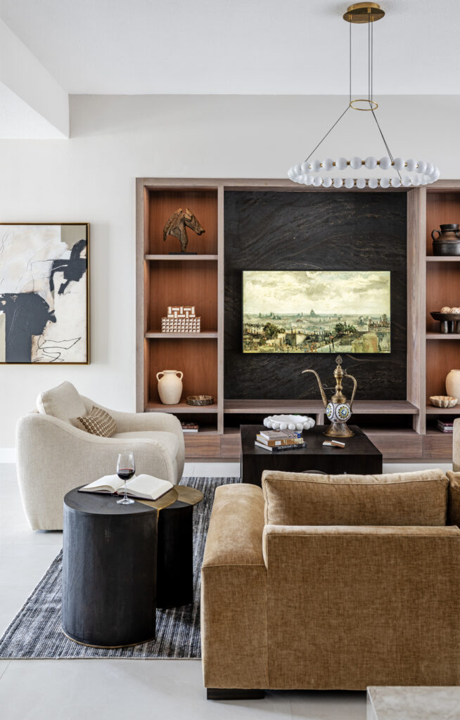 Modern warm custom entertainment center in Bradenton FL. Warm living room featuring a custom wood and black stone built-in entertainment wall, cream curved accent chair, caramel sectional, dark drum side tables, and a beaded globe chandelier — RDM Interiors