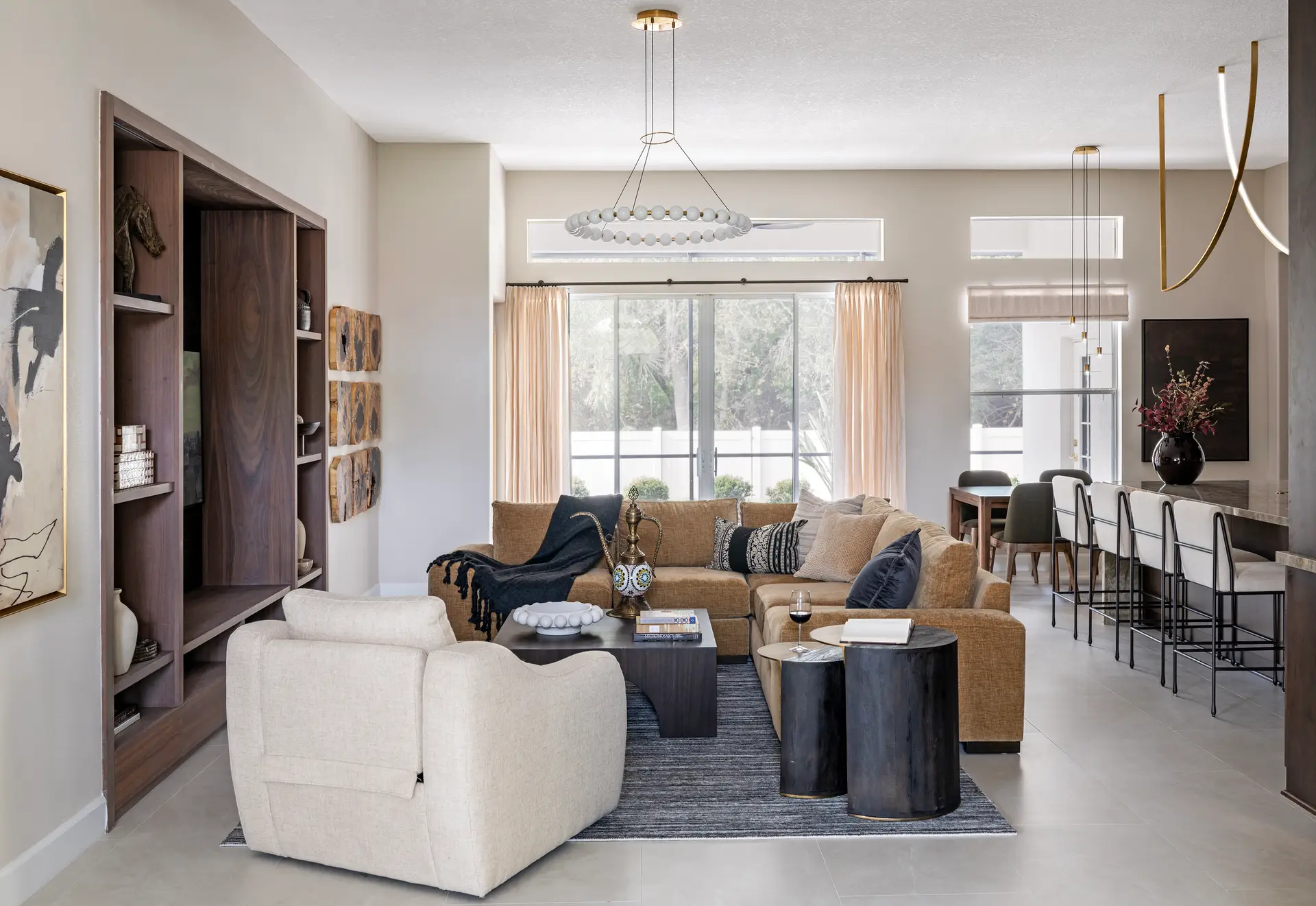 Open-concept living and dining space with a caramel sectional, cream accent chair, dark wood built-in shelving, a beaded chandelier, and a gold sculptural pendant over the dining area — RDM Interiors
